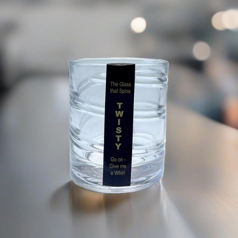 A clear glass tumbler with a black vertical label that reads 'The Glass that Spirs TWISTY Go on - Give me a Whirl' on a blurred indoor tabletop background.