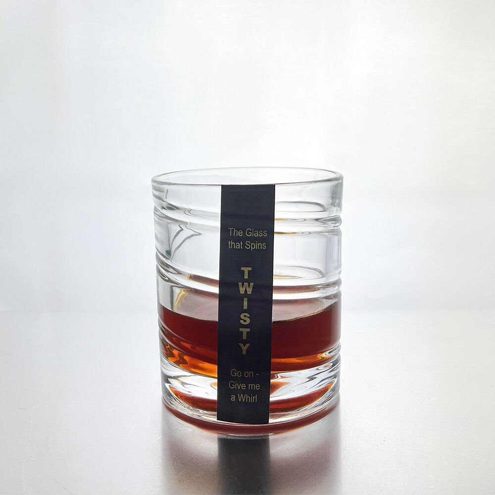 A clear glass rocks tumbler with horizontal ridges, filled with an amber-brown liquid, featuring a black vertical label that reads 'TWISTY' with the tagline 'The glass that Spins Go on - Give me a Whirl'.