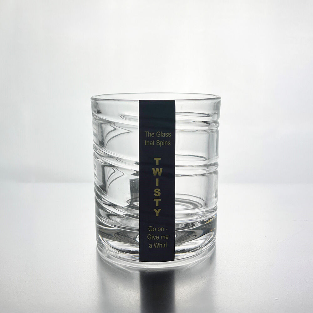 A clear glass rocks tumbler with horizontal ridges and a black vertical label that reads 'TWISTY' with smaller text above and below, photographed on a light gray reflective surface.