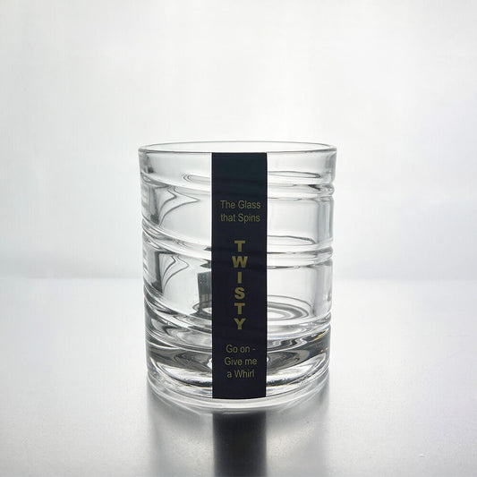 A clear glass rocks tumbler with horizontal ridges and a black vertical label that reads 'TWISTY' with smaller text above and below, photographed on a light gray reflective surface.