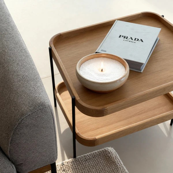 Wooden side table with a candle and Prada book next to a gray sofa.