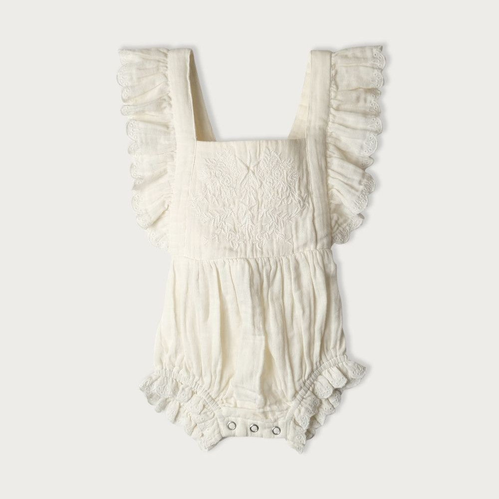 A white bodysuit with embroidered detailing on the front panel and ruffles, featuring an adjustable tie up back and an elastic waistband for babies.