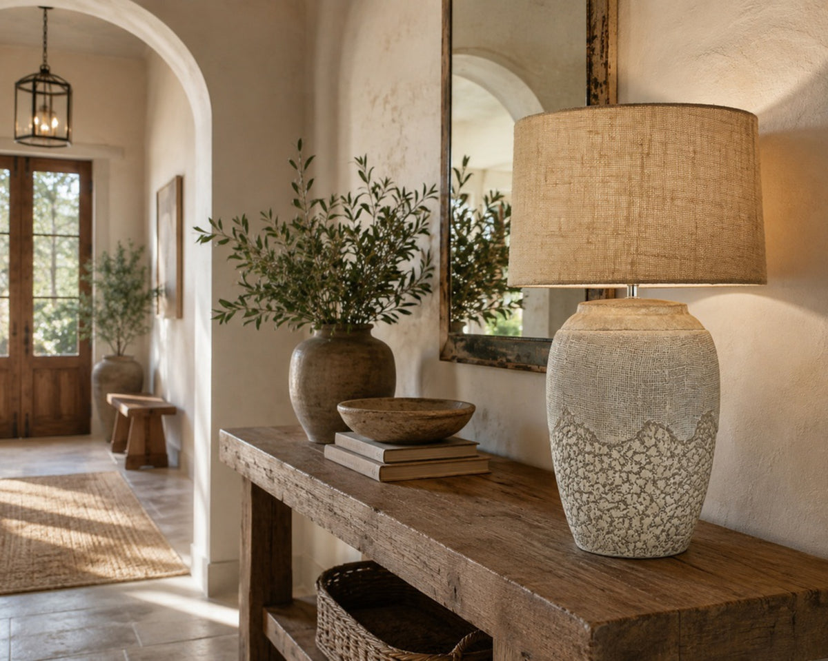 A rustic ceramic table lamp with a textured beige base and a natural linen drum shade, styled on a wooden console table in a warm, sunlit home interior.