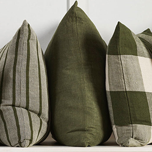 Five decorative throw pillows in various patterns and colors, including solid olive green, striped green and beige, and plaid green and cream, arranged on a light wood shelf against a white paneled wall.